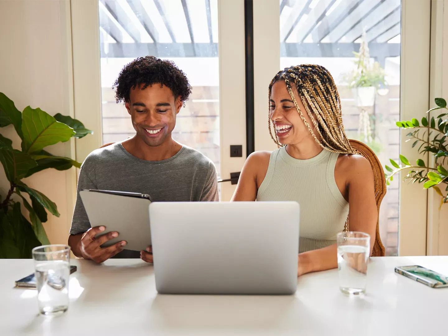 Smiling couple, female using laptop and male using a tablet, both enjoying the internet in their naturally lit kitchen.