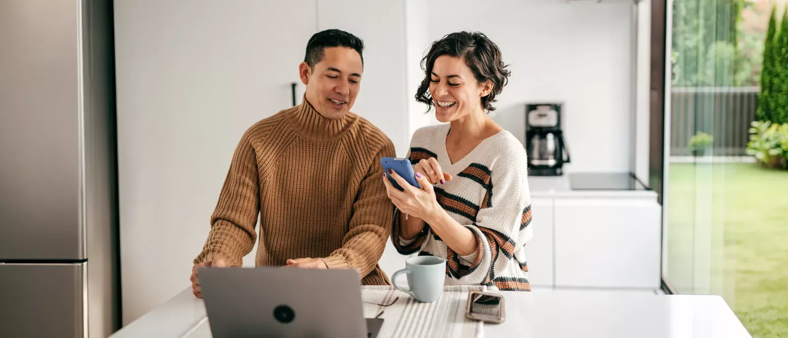 Smiling couple using a laptop and smartphone inside their kitchen.