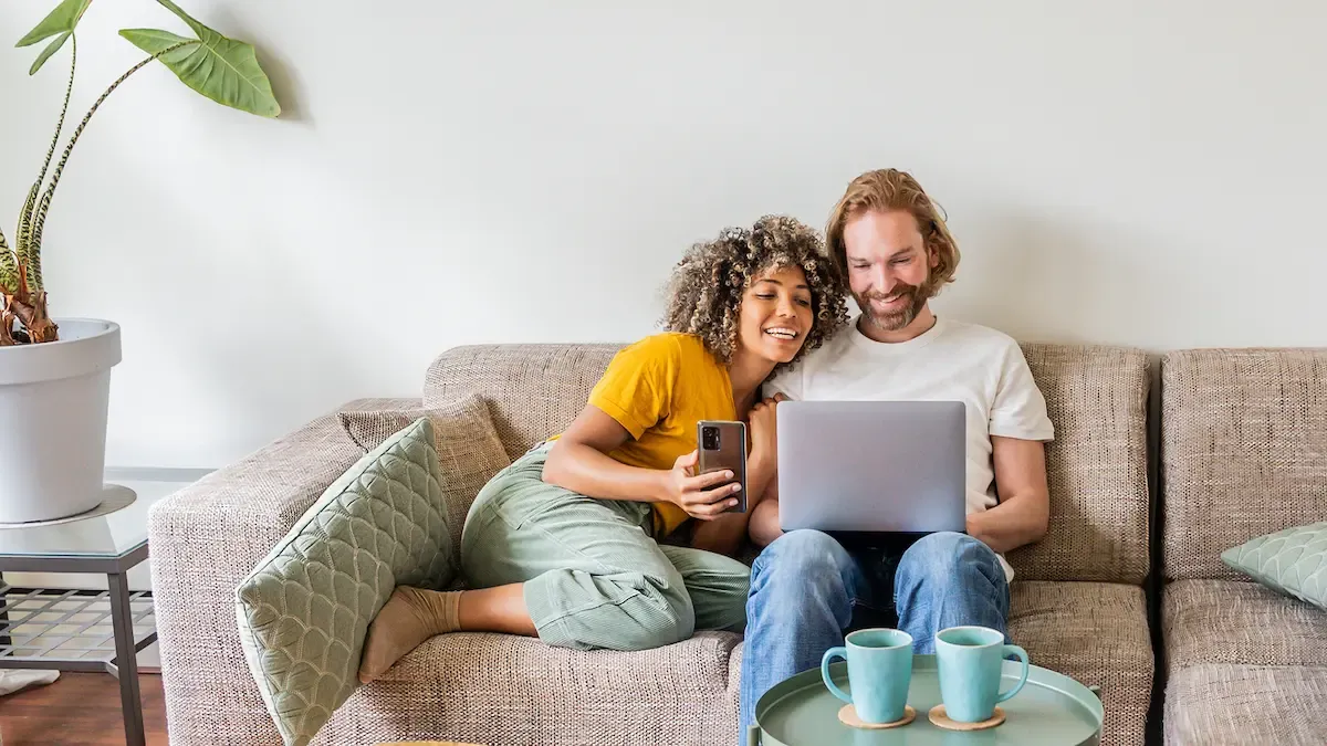 A smiling couple video chats together using a phone and laptop on the couch.
