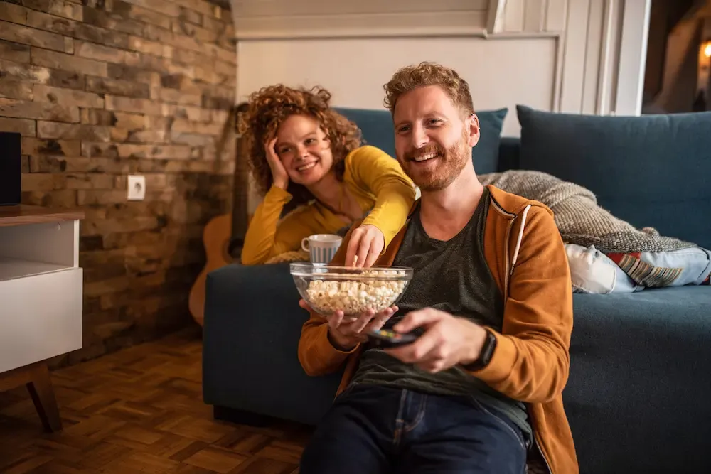 A smiling couple enjoys popcorn and watches television while relaxing in a cozy living room.