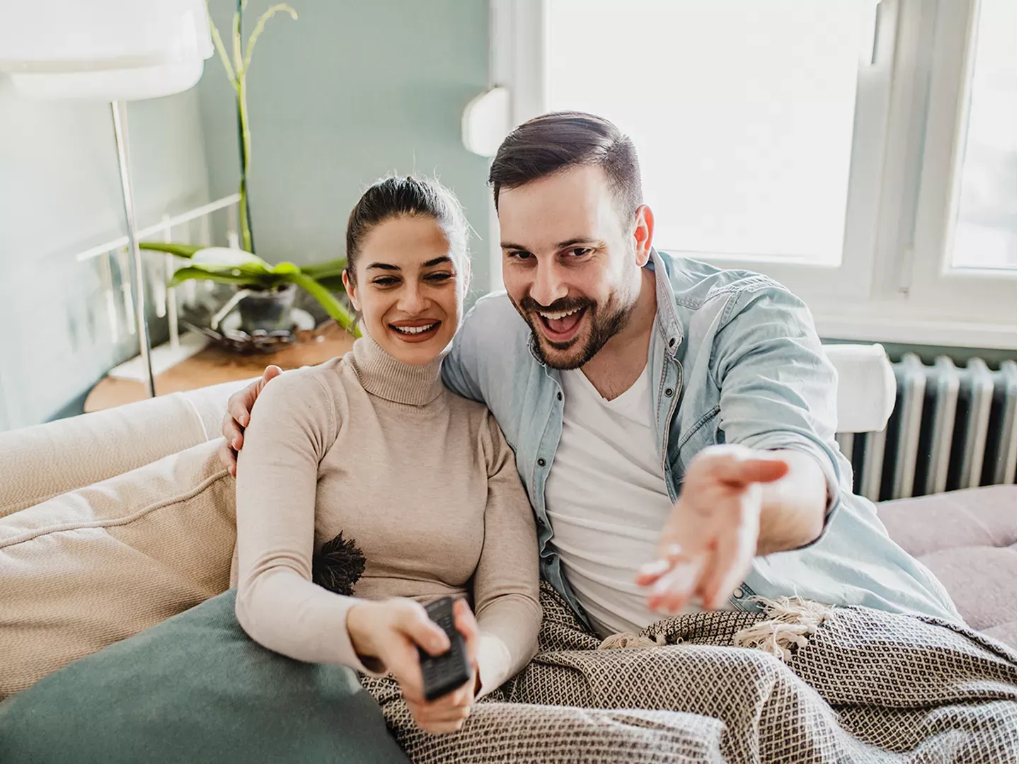 A happy couple sits close together on a couch while using a remote to watch television.