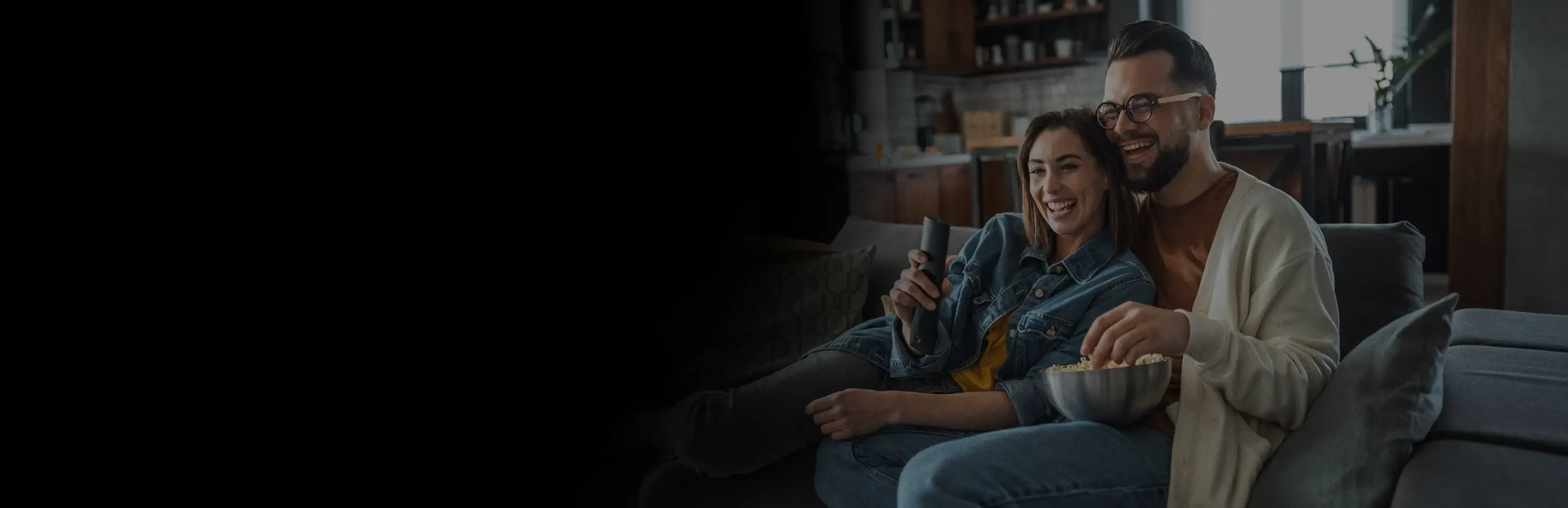 A laughing couple cuddles on a gray sofa, sharing a bowl of popcorn and holding a remote control.