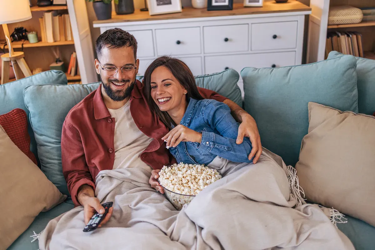 A smiling couple cuddles under a blanket sharing popcorn while watching TV.