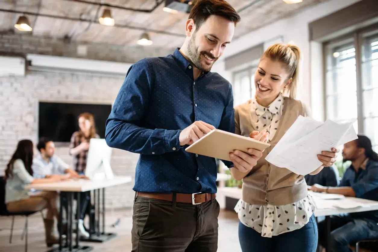 Two smiling colleagues stand in a modern, open-plan office discussing work while looking at a digital tablet and paper documents.