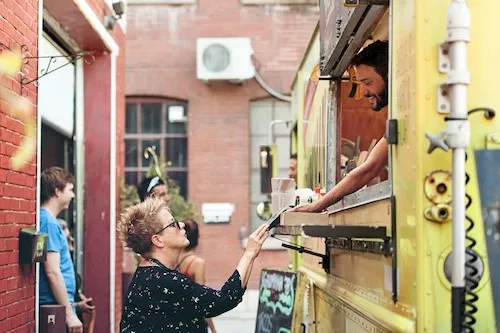 A customer with short blonde hair is handing a menu to a smiling vendor inside a bright yellow food truck parked in an urban alley.