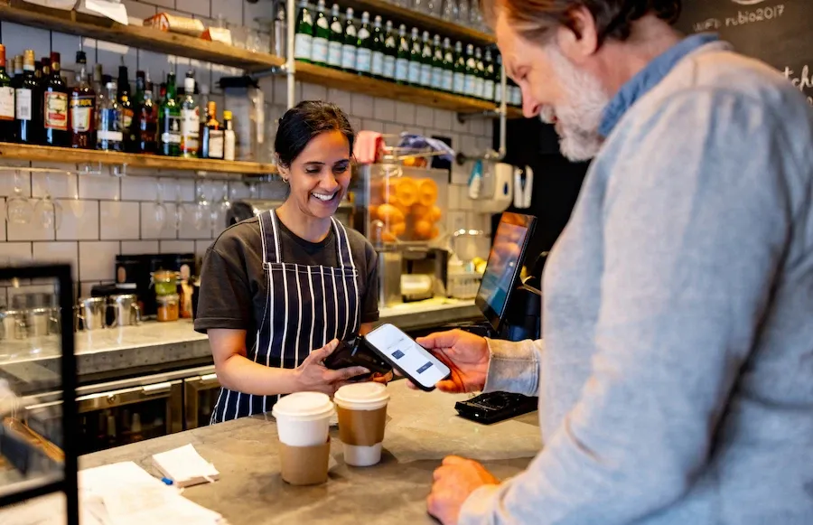 A smiling female barista holds a payment terminal while a customer pays for his coffee order using contactless payment on his smartphone.