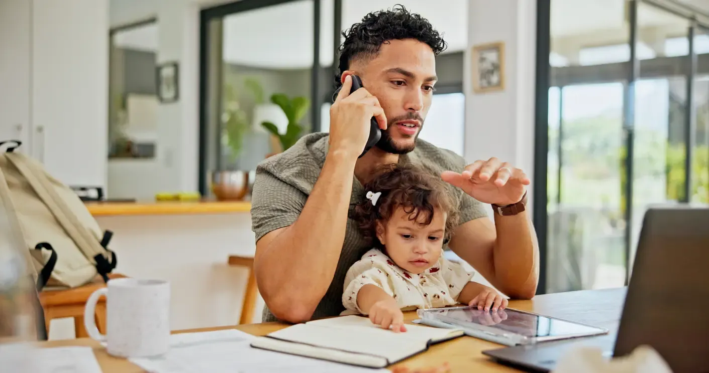 A father takes a phone call while holding his toddler at a busy home desk.