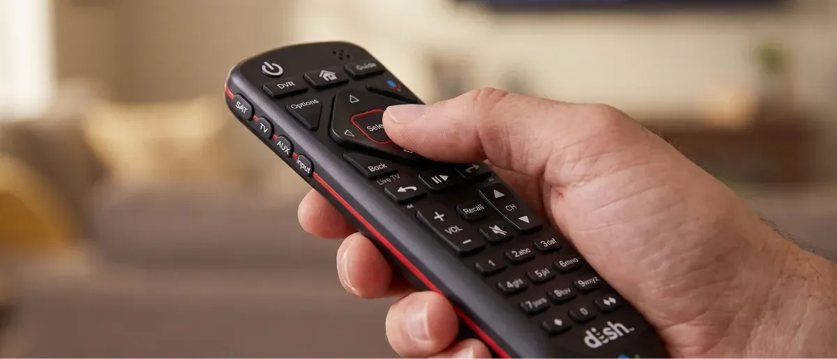 A hand holds a black DISH remote pointed at a blurred television.