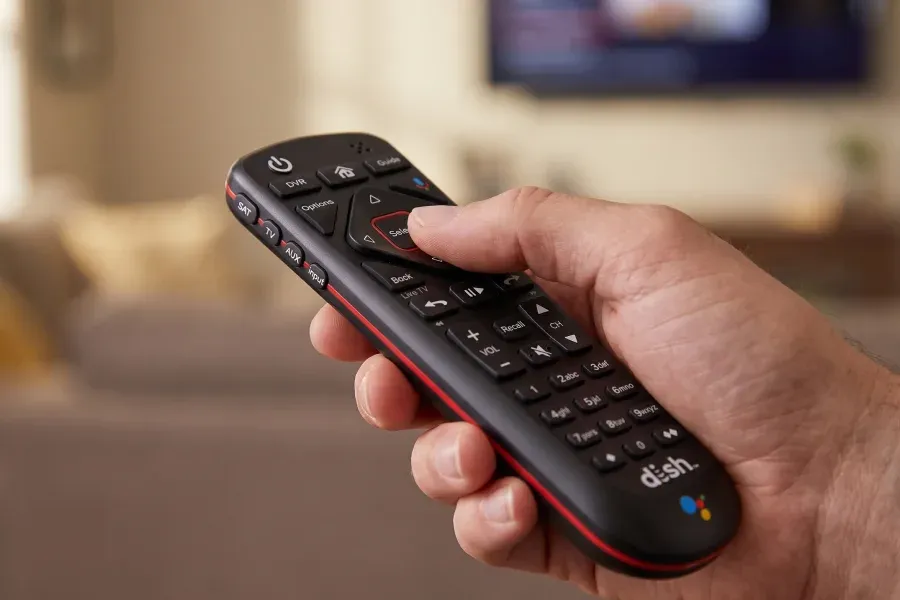 A hand holds a black DISH remote pointed at a blurred television.