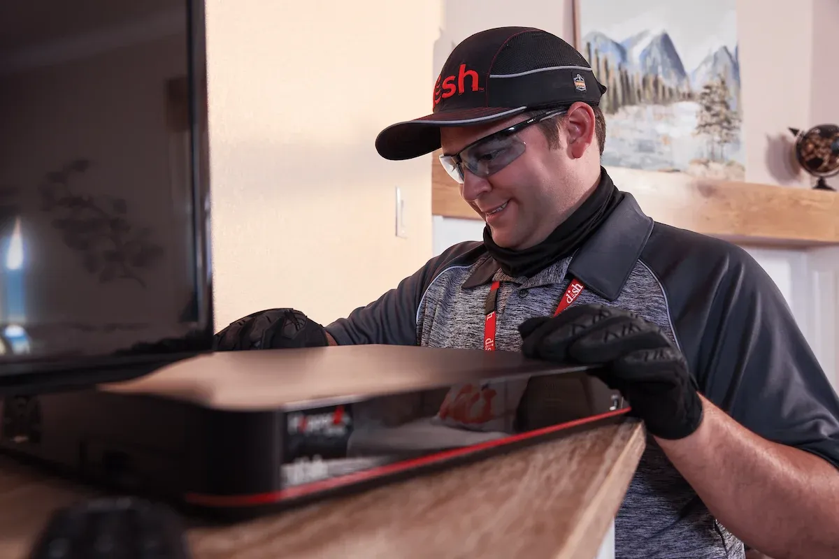 A DISH technician in safety glasses installs a Hopper receiver on a wooden entertainment stand.