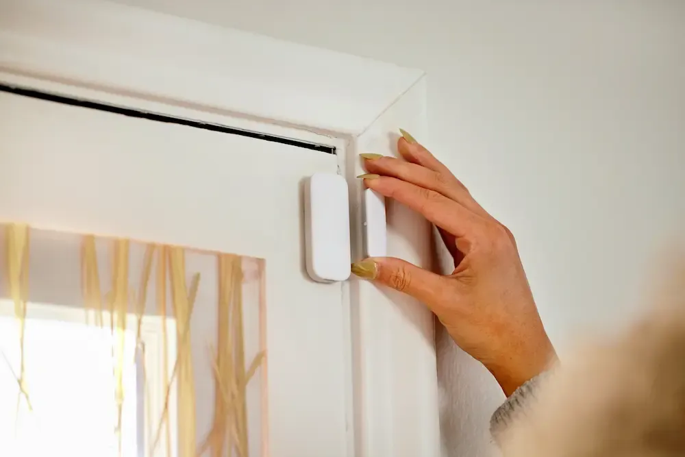 A person's hand installing a white DIY door and window security sensor onto a white door frame.