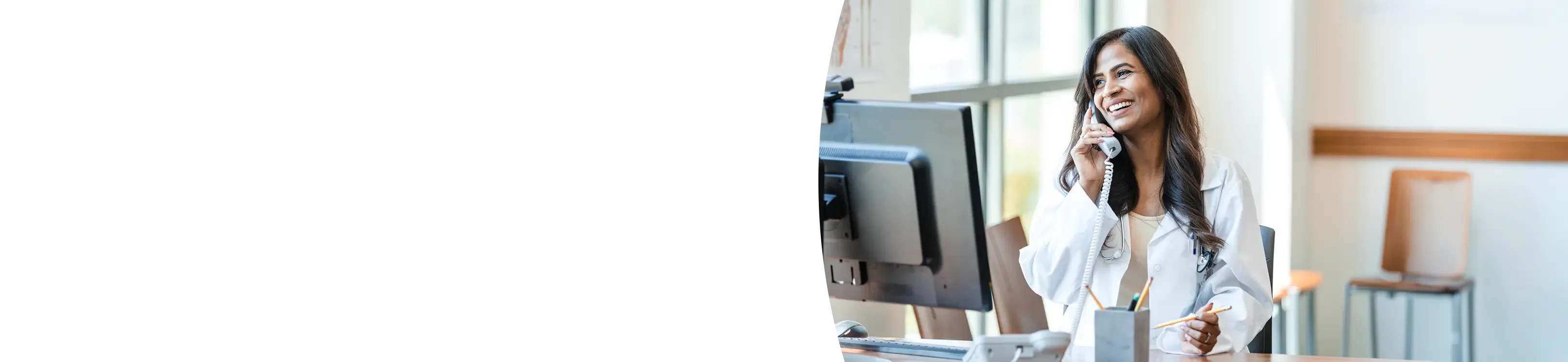 A smiling female doctor or medical professional wearing a white lab coat is sitting at a desk and talking on a landline phone next to a computer monitor.