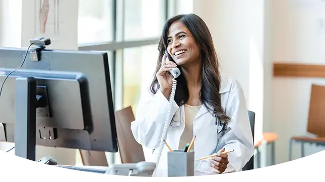 A smiling female doctor or medical professional wearing a white lab coat is sitting at a desk and talking on a landline phone next to a computer monitor.