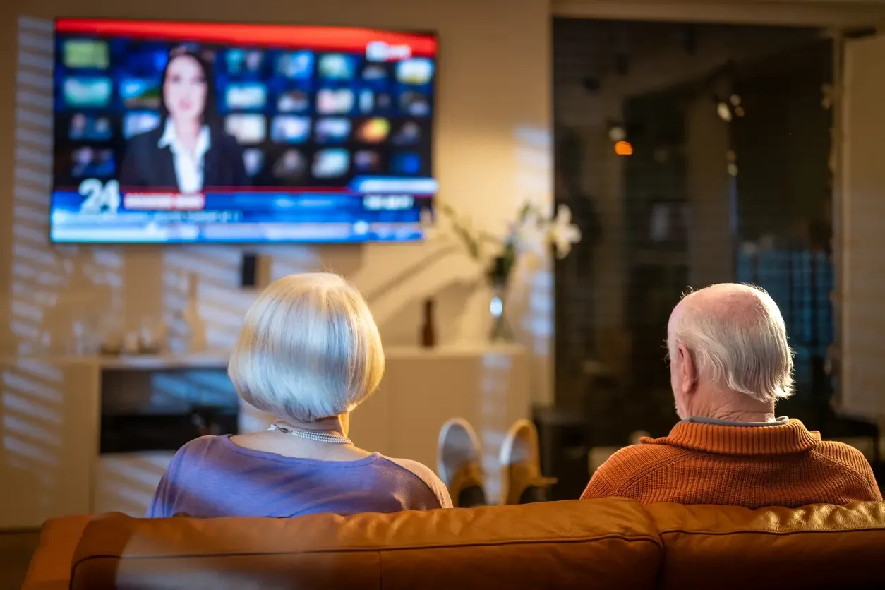 An elderly couple sits on a leather sofa watching a news broadcast on a large wall-mounted TV.