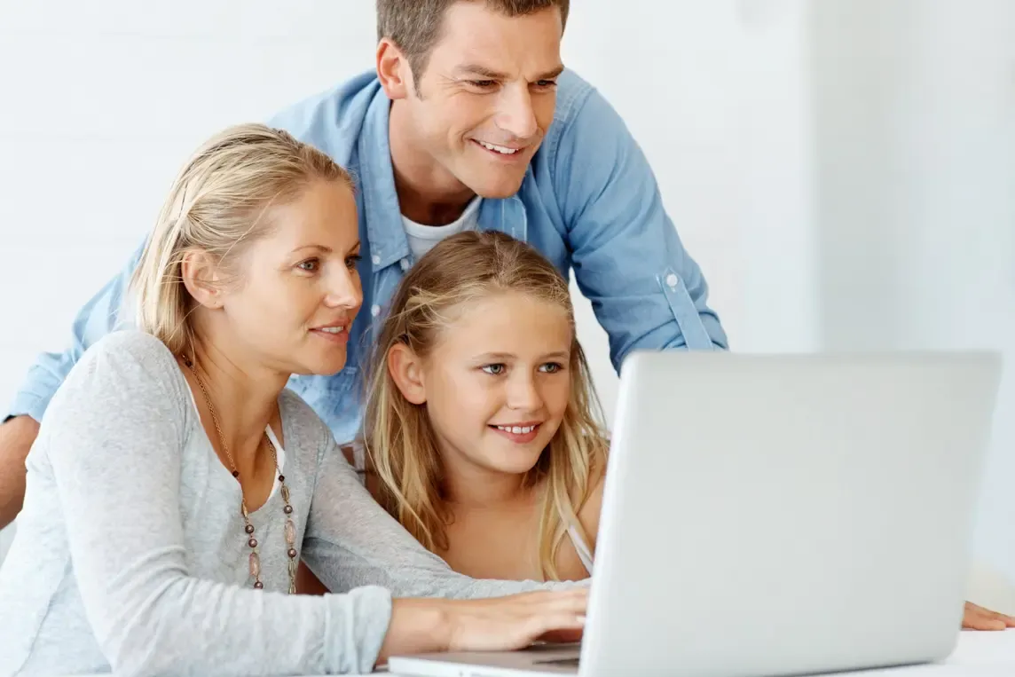 A smiling mother, father, and young daughter sitting together and looking at a laptop screen.