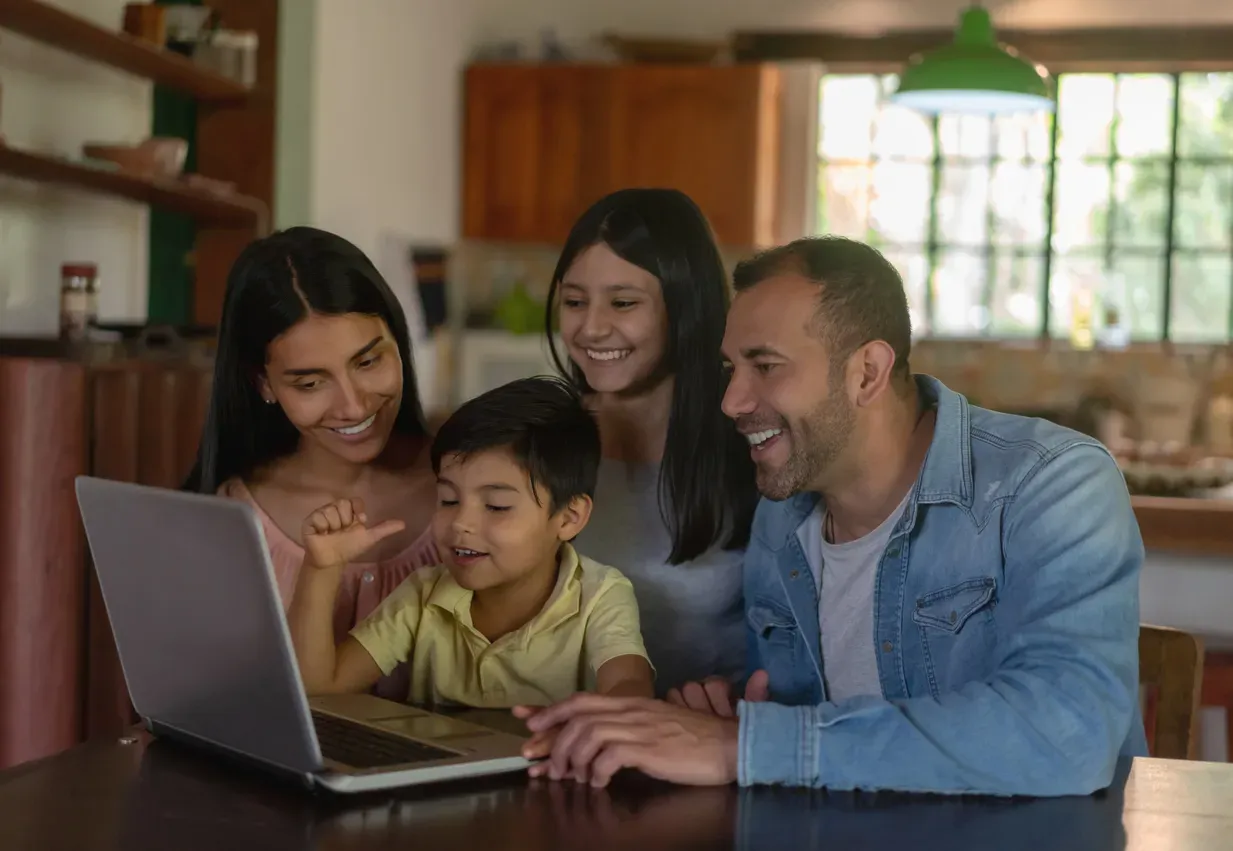 A smiling family of four gathers around a laptop at a kitchen table.
