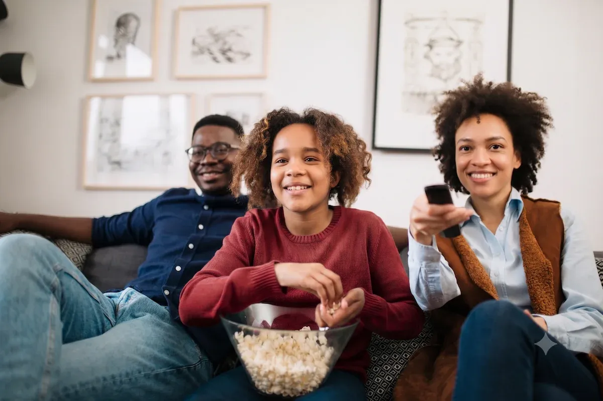 A family sits on a couch sharing popcorn while holding the TV remote.