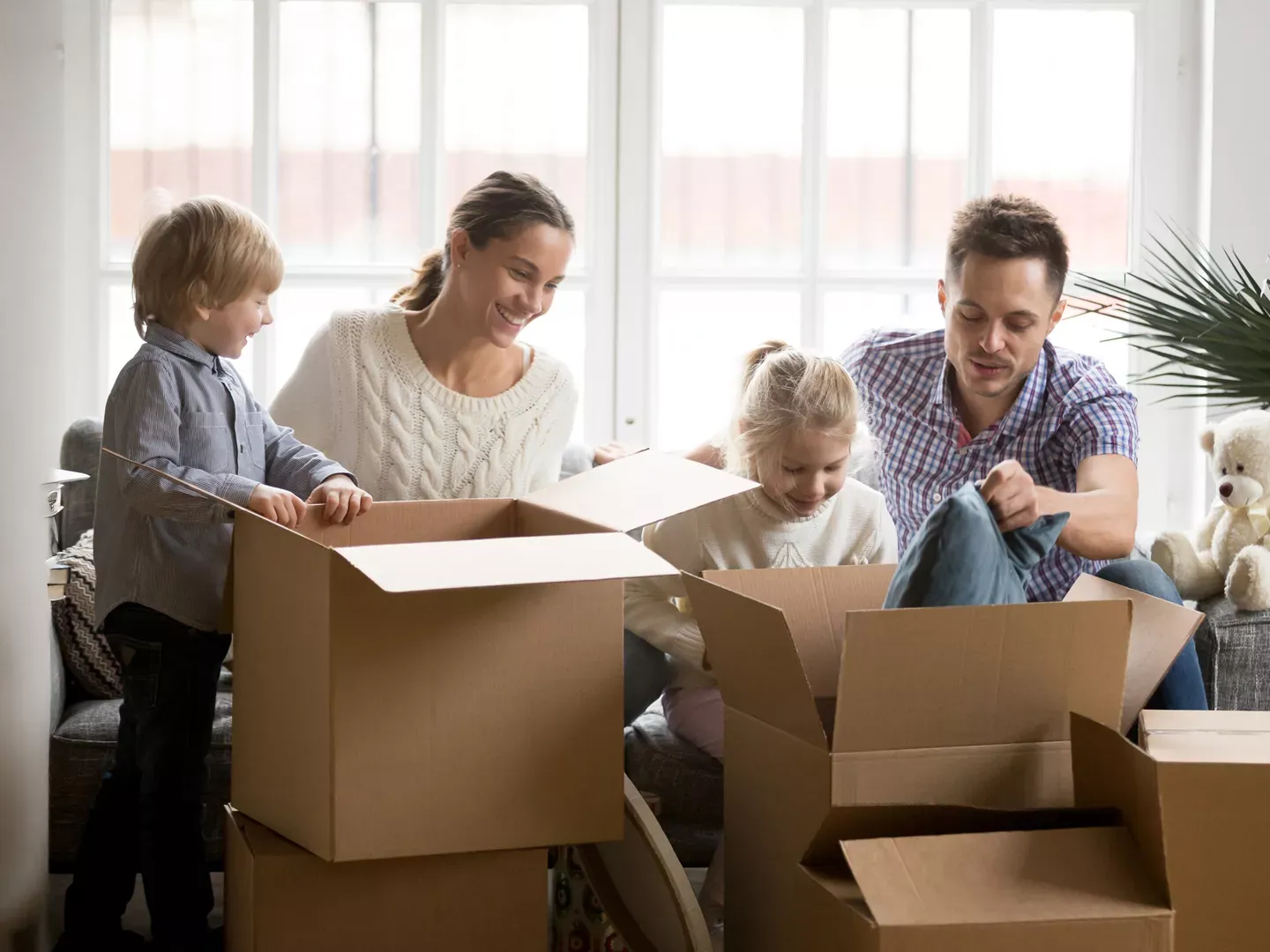 A happy family of four gathers in their living room to unpack cardboard boxes together.
