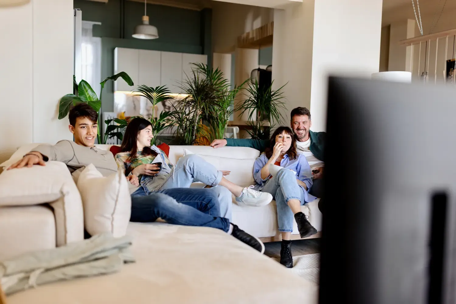 A family of four sits together on a large white sectional sofa watching a movie in a modern, open-concept home.