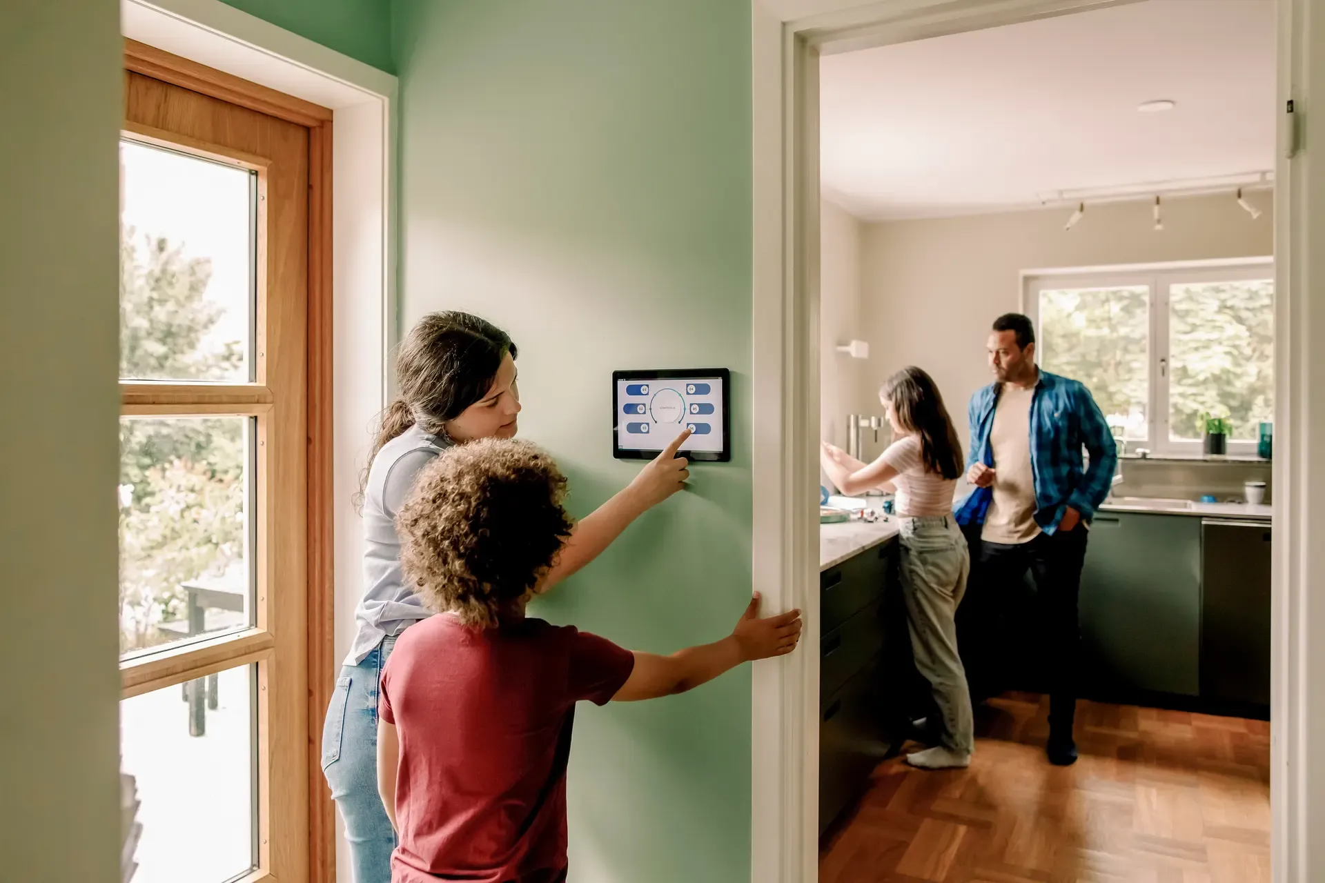 A woman and young boy use a wall-mounted smart home control panel while a man and girl stand in the kitchen in the background.