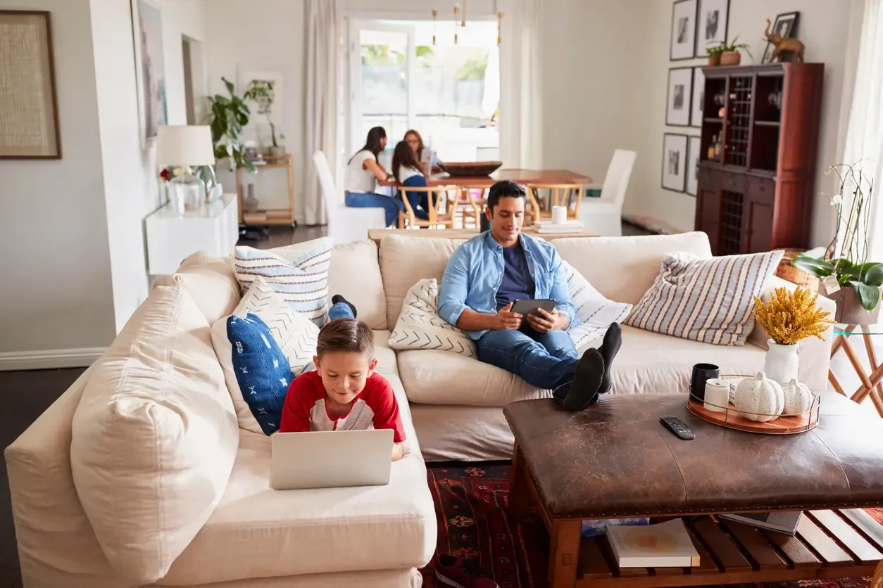 A boy uses a laptop while his father reads a tablet on a sectional sofa at home.