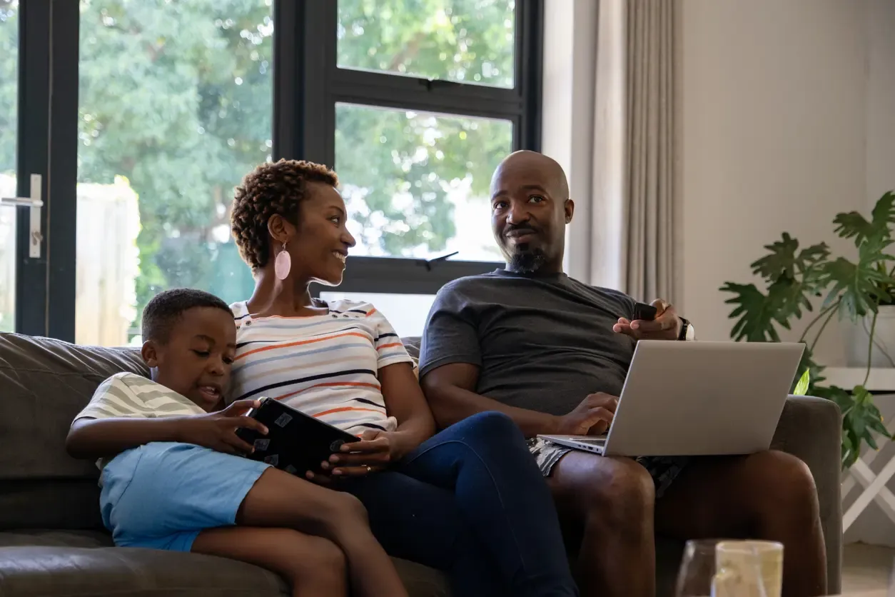 A family of three sitting on a living room sofa while using a laptop and a tablet together.