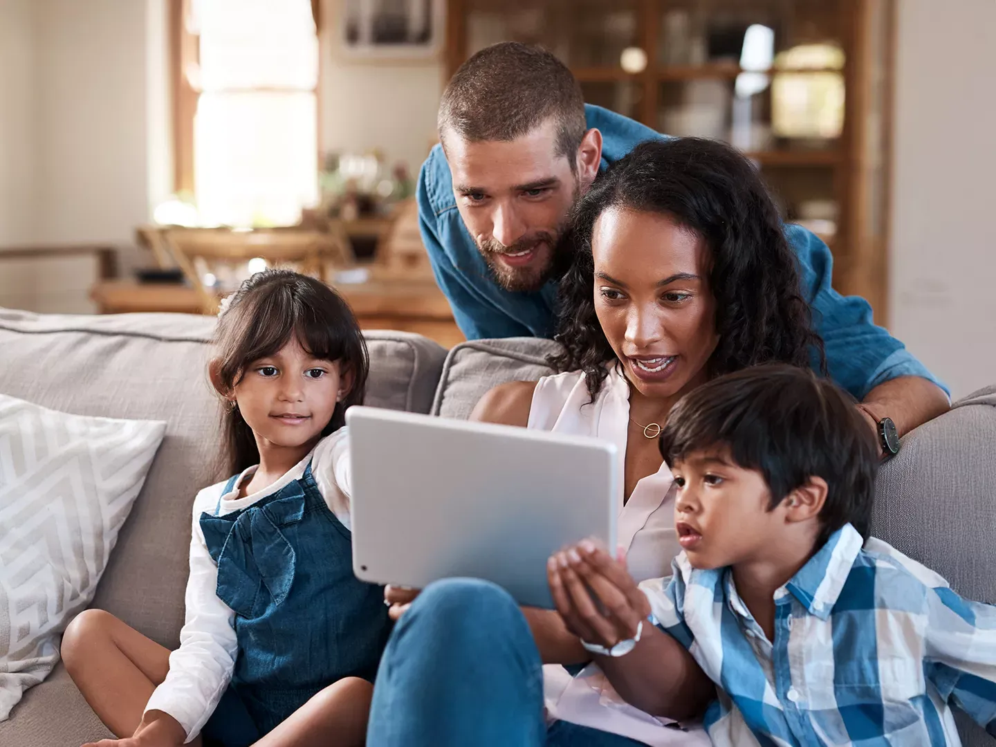 A father, mother, and two young children gather around a tablet while sitting on a living room sofa.
