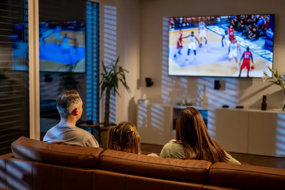 A family of three sits on a brown leather couch in a dimly lit living room while watching a basketball game on a large television.