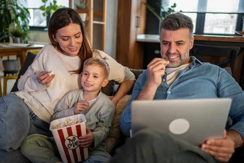 A mother, father, and young son sitting on a couch together eating popcorn and smiling while watching a video on a laptop.