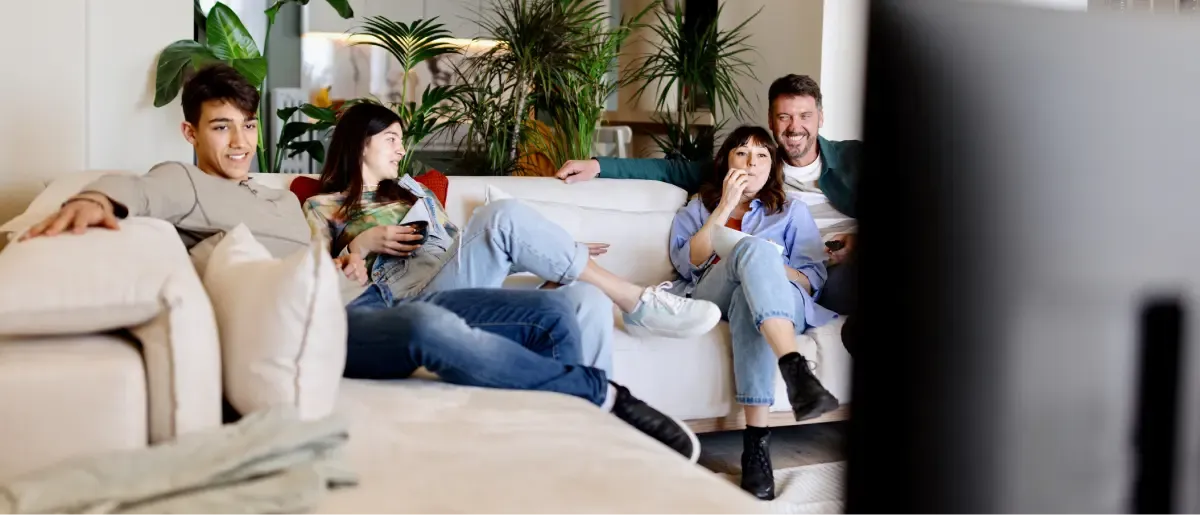 A family of four relaxes on a white couch watching television in a plant-filled living room.