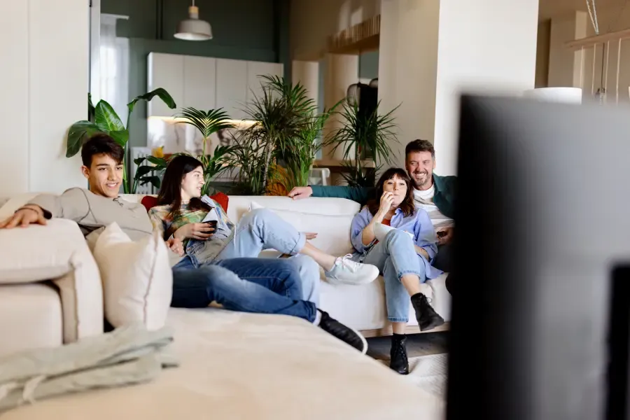 A family of four relaxes on a white couch watching television in a plant-filled living room.
