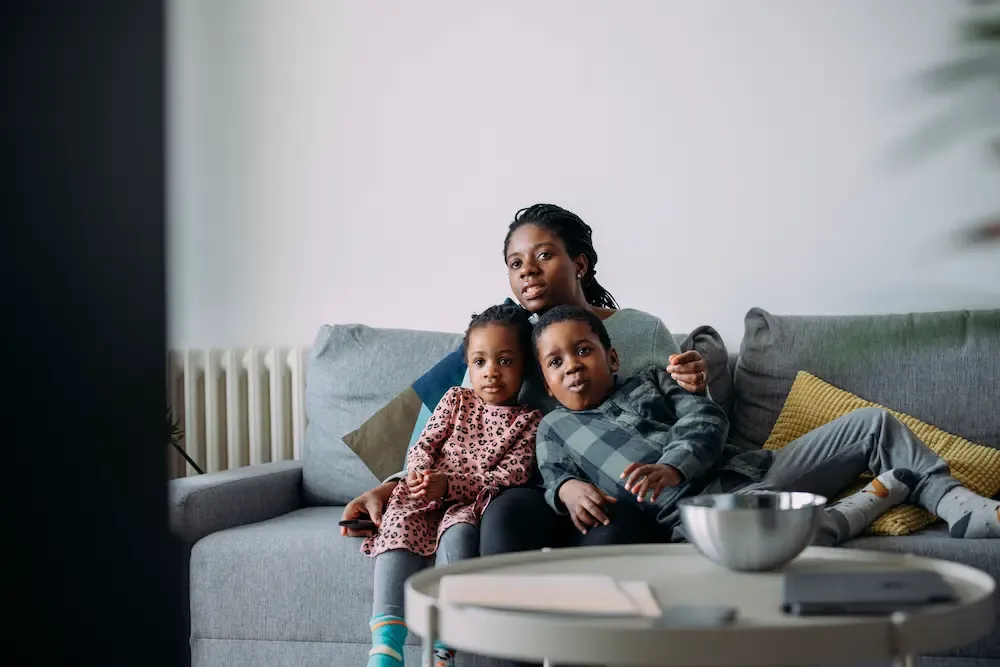 A mother sits on a gray sofa with her two young children, all looking toward a television screen in a bright living room.