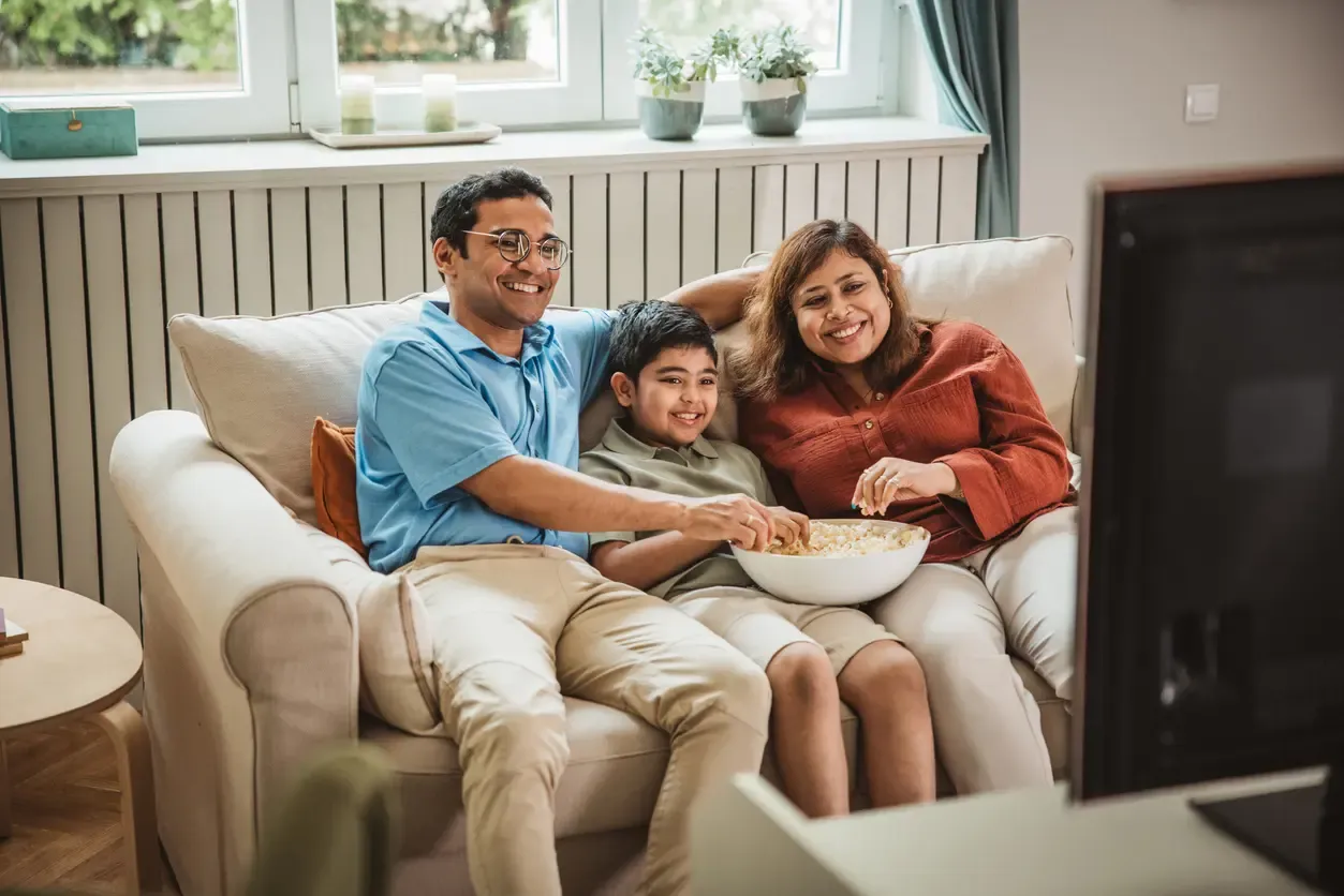 A smiling family of three shares a bowl of popcorn on a couch while watching TV together.