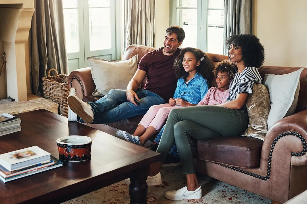 A diverse family, including a father, mother, and two young daughters, sit smiling together on a comfortable brown leather sofa, watching television in a sunlit living room.