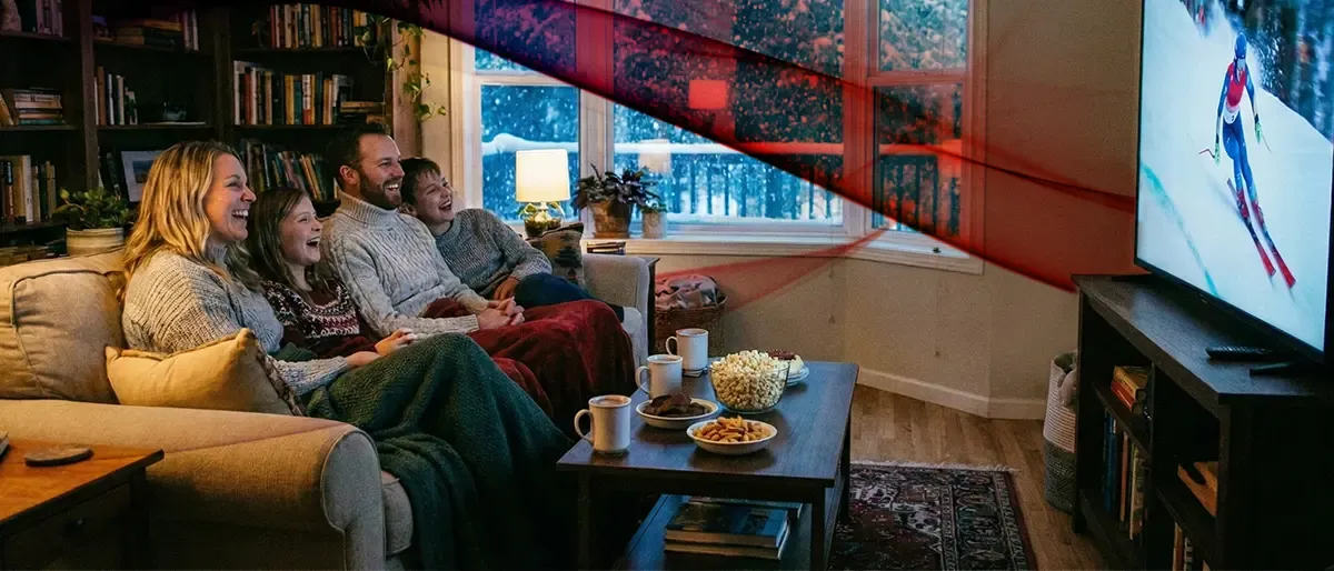 A happy family of four sits on a couch under blankets, laughing while watching a downhill skier on television in a cozy living room during a snowstorm.