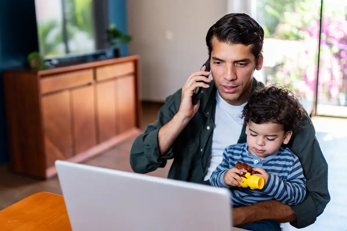A man talks on the phone at a laptop while holding a toddler with a toy on his lap.