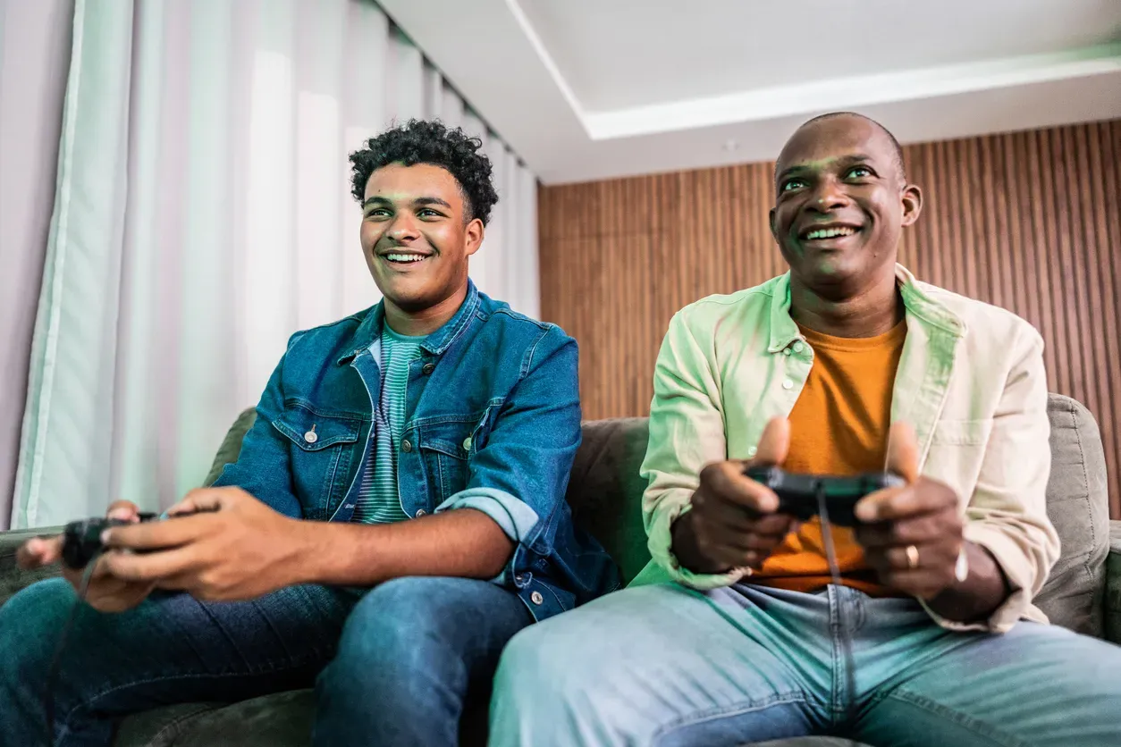 A father and teenage son smile while playing video games with controllers on a couch.