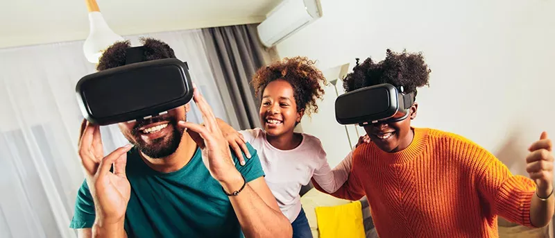 A father, mother, and child smiling and having fun while wearing virtual reality headsets in their living room.