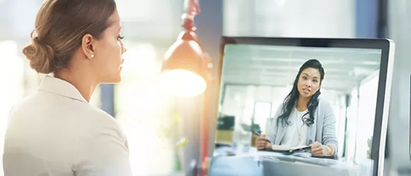 A woman sitting at a desk has a video call with another woman displayed on her computer monitor.