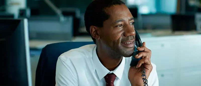 A professional man in a white shirt and tie smiles while speaking on a black corded office phone.
