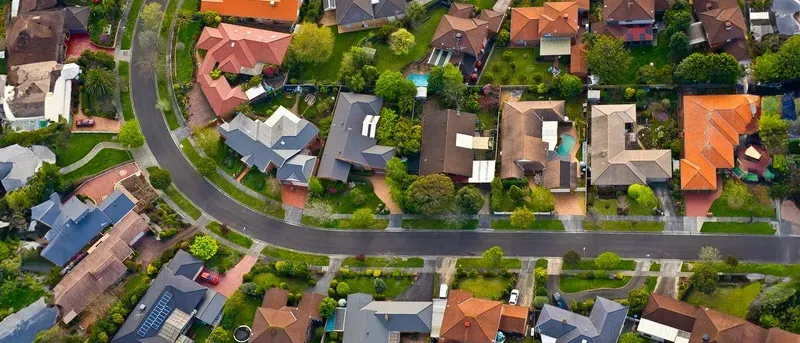 An aerial view of a densely populated suburban neighborhood with curving streets, green yards, and houses with various colored roofs.