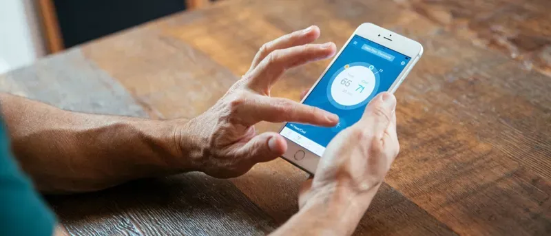 A close-up of a person's hands using a smartphone to adjust a smart thermostat application on a wooden table.
