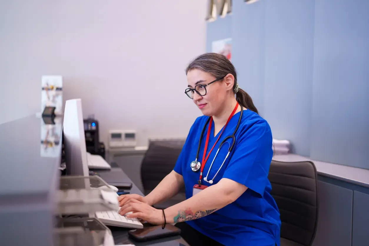 A female healthcare professional in blue scrubs with a stethoscope around her neck typing on a computer at a clinic reception desk.