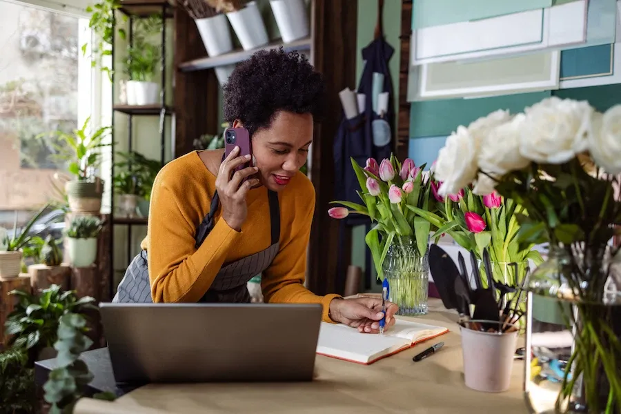 A smiling Black woman in a mustard yellow top and apron is talking on a smartphone and writing in a notebook behind a counter in a flower shop.