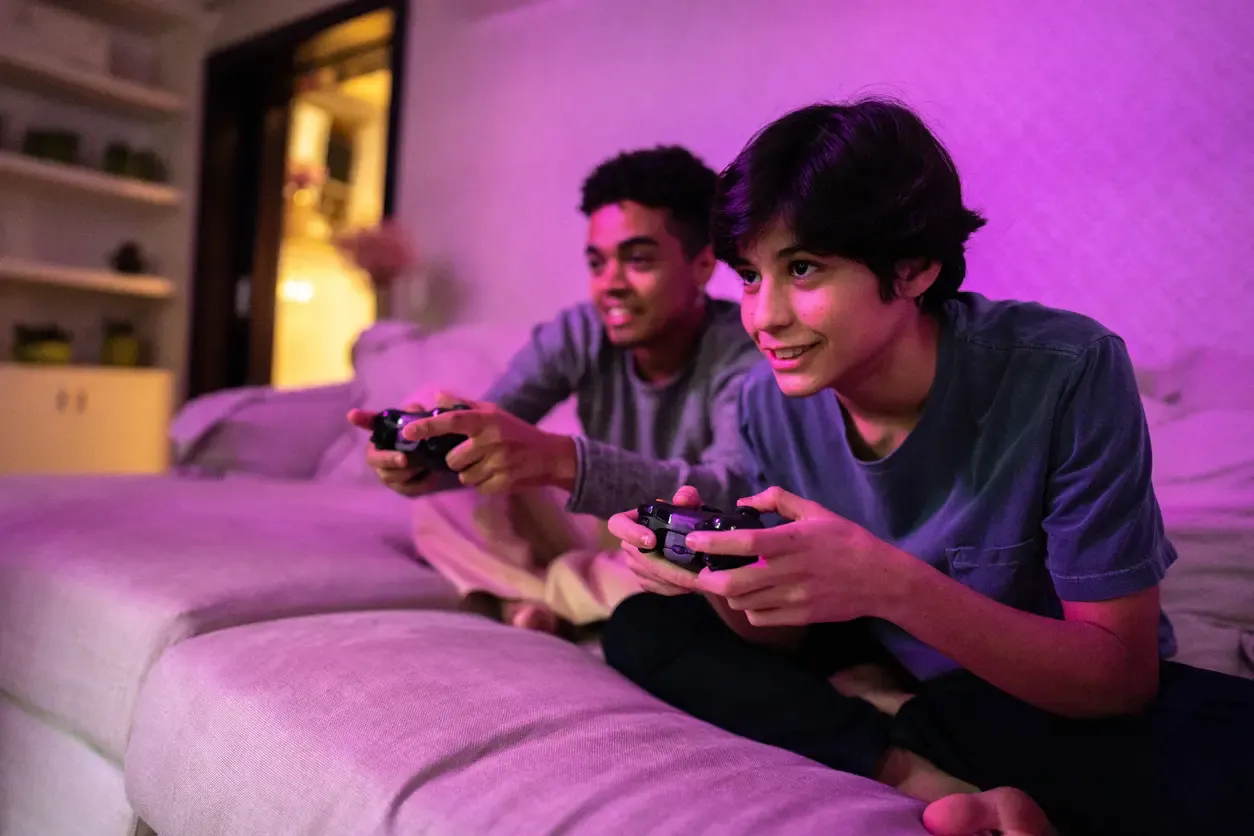 Two young people sit together on a sofa as one holds a game controller and they watch a screen intently.