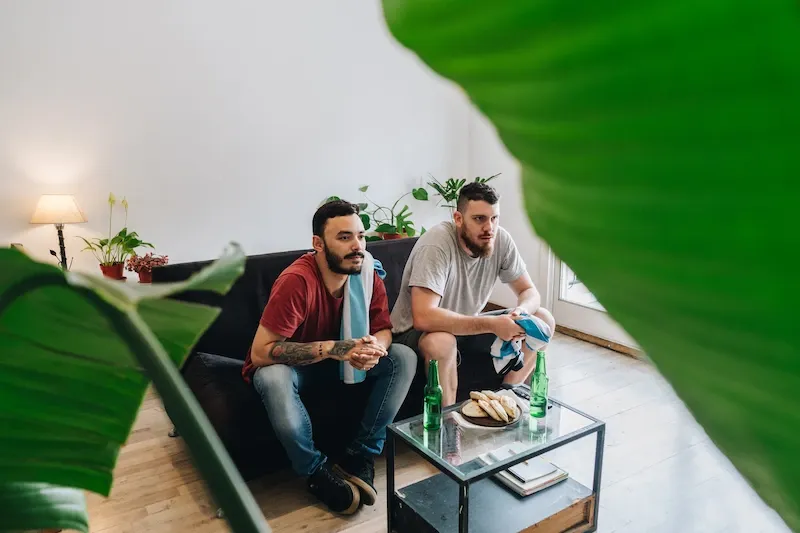 Two friends sit on a sofa watching a sports match with an Argentina flag, beer, and snacks on the coffee table.