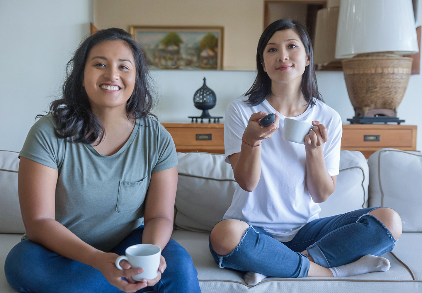 Two roommates binge-watching their favorite TV series. They are sitting on a sofa and holding coffee mugs.