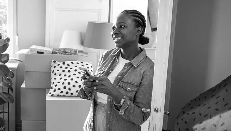 A smiling woman standing in a bright living room filled with moving boxes while holding a smartphone.