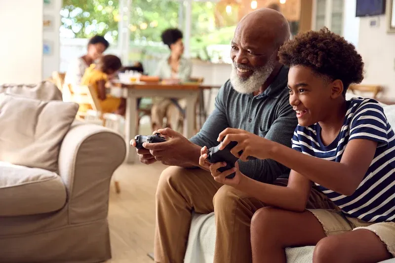 A smiling grandfather and grandson play video games together on a living room couch, with other family members gathered at a table in the background.