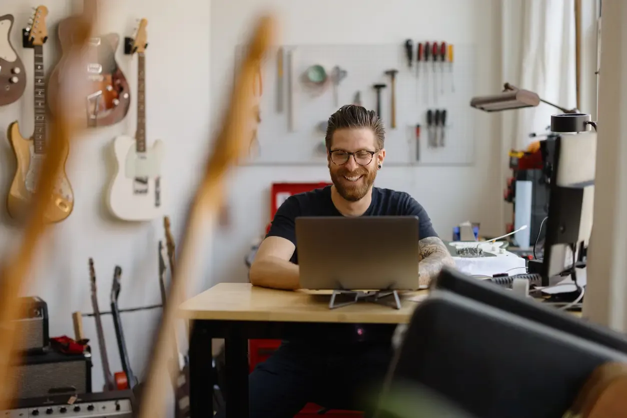 A smiling man works on a laptop in a guitar repair workshop surrounded by hanging instruments and tools.